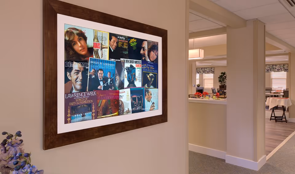 A framed collage of vintage album covers hanging on a beige wall in an interior space. To the right, there is a partial view of a room with tables covered in white tablecloths and chairs, suggesting a dining area. The space is softly lit with ceiling lights and has a warm, inviting atmosphere.