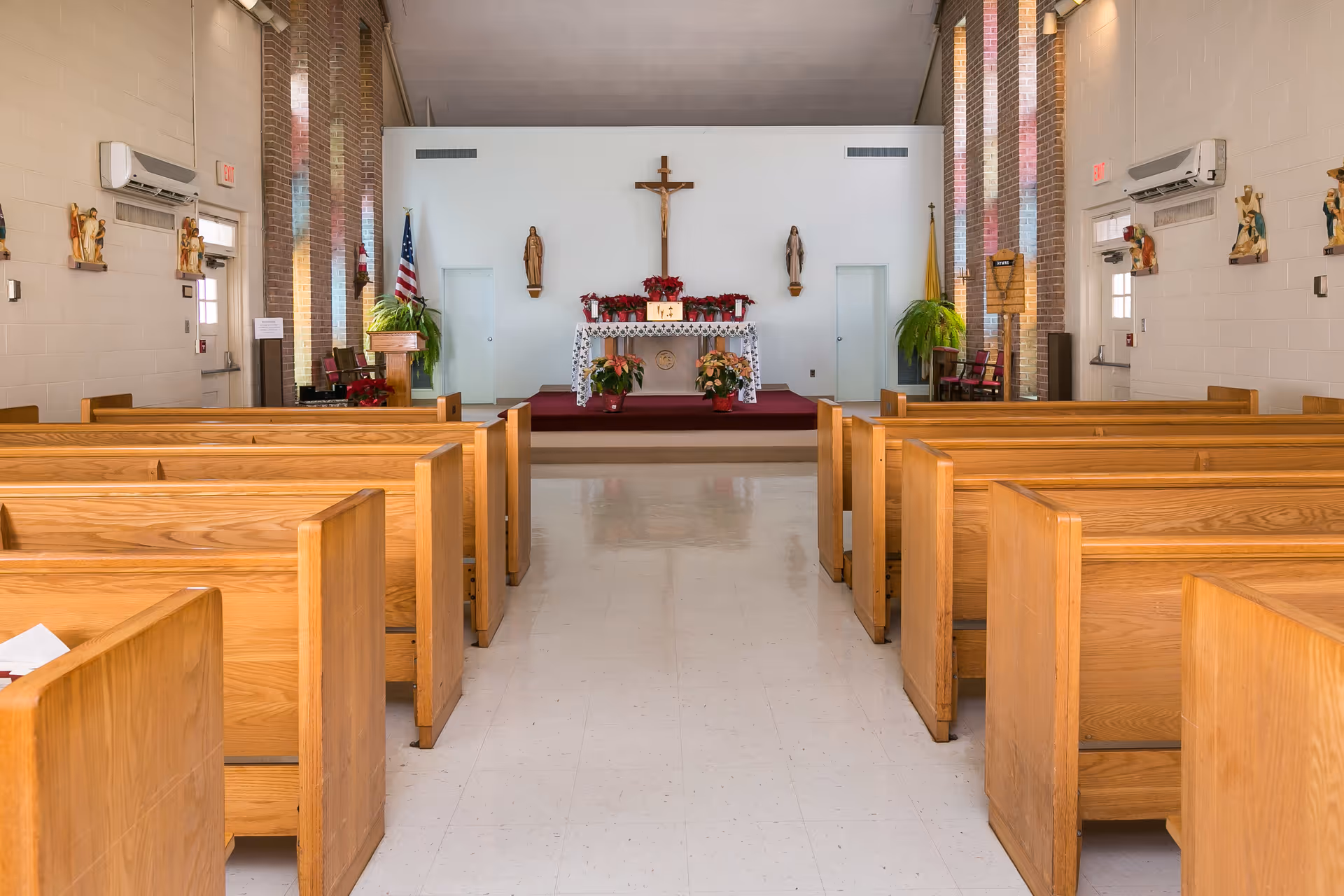 Interior view of a chapel with wooden pews arranged in rows facing an altar. The altar is decorated with flowers and religious statues, including a crucifix mounted on the wall behind it. The chapel has brick columns, white walls, and windows letting in natural light.