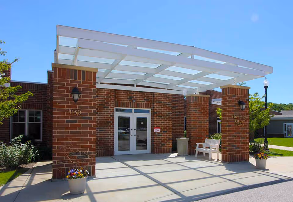 Brick-faced building entrance with a white overhead pergola, double glass doors, potted flowers and a bench.