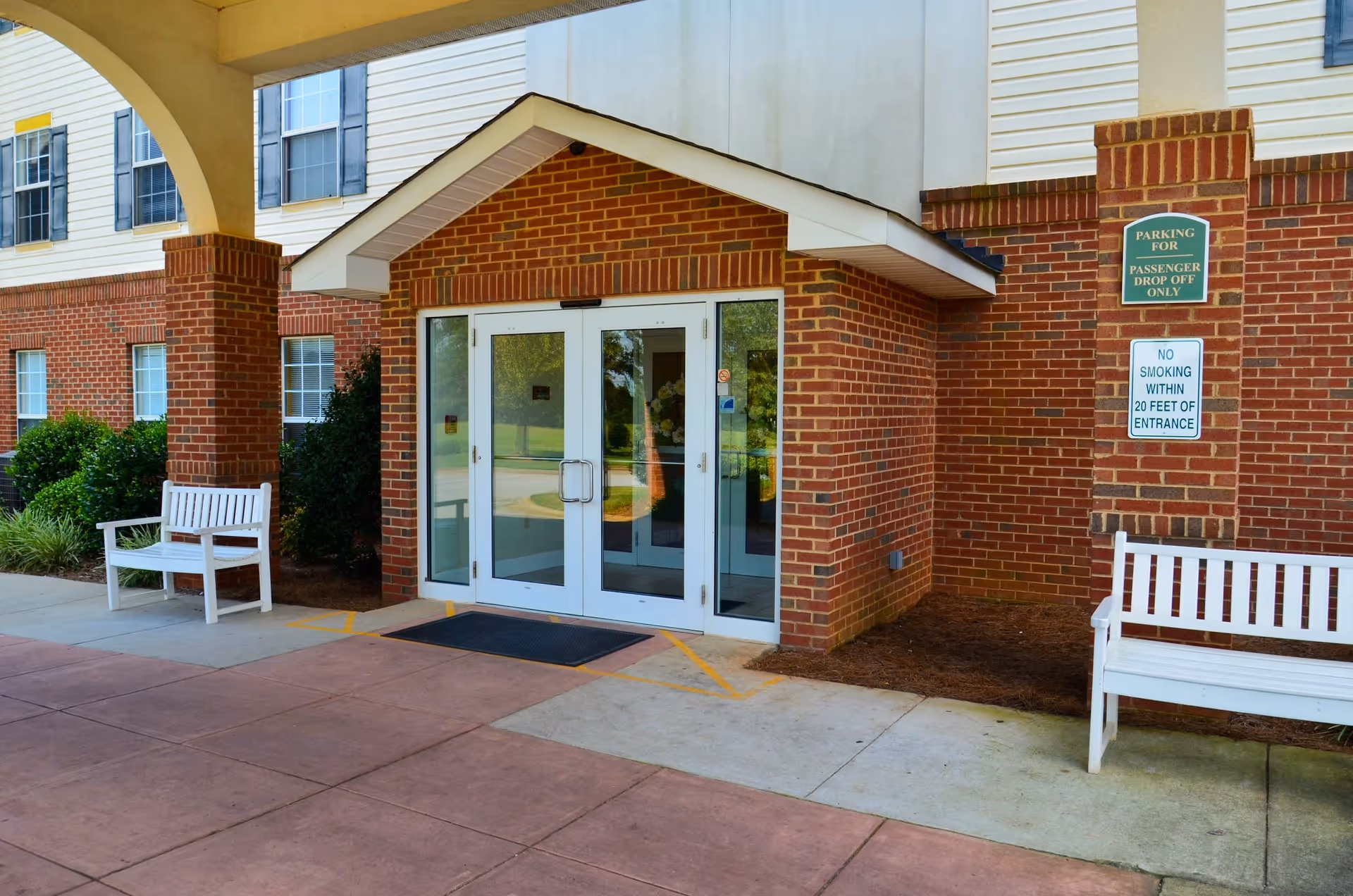 Entrance to a brick building with double glass doors under a covered walkway. There are two white benches on either side of the entrance and signs indicating parking for passenger drop off only and no smoking within 20 feet of the entrance.