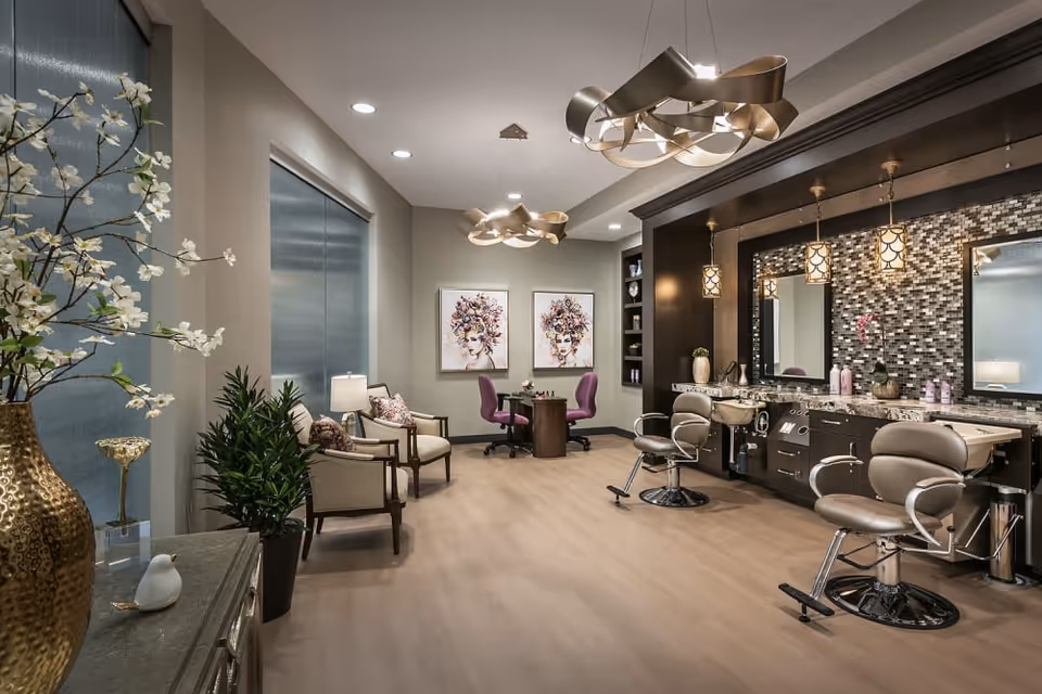 A modern salon area in a senior living facility featuring two styling chairs in front of a large mirror with mosaic tile backsplash, pendant lights, and a countertop with hair care products. The room also has a small seating area with two armchairs and a table with two purple chairs, decorative artwork on the wall, and plants for ambiance.