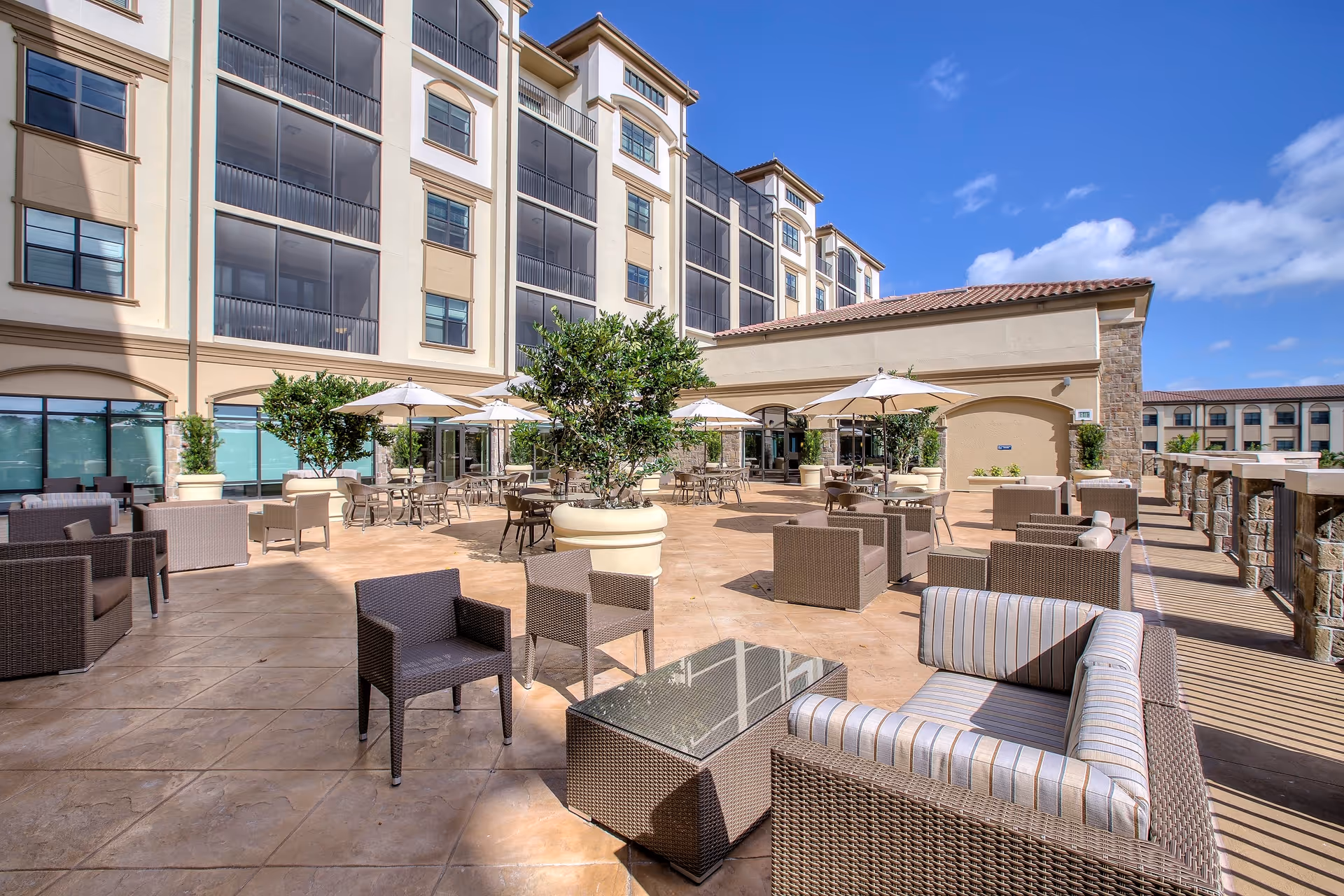 Outdoor patio area at The Arlington of Naples featuring multiple seating arrangements with wicker chairs, sofas, glass-top tables, and umbrellas. The patio is spacious with large potted plants and is adjacent to a multi-story building under a partly cloudy blue sky.
