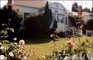 Exterior view of a single-story residential building with a small front porch, surrounded by greenery and flowering plants, under a clear sky.