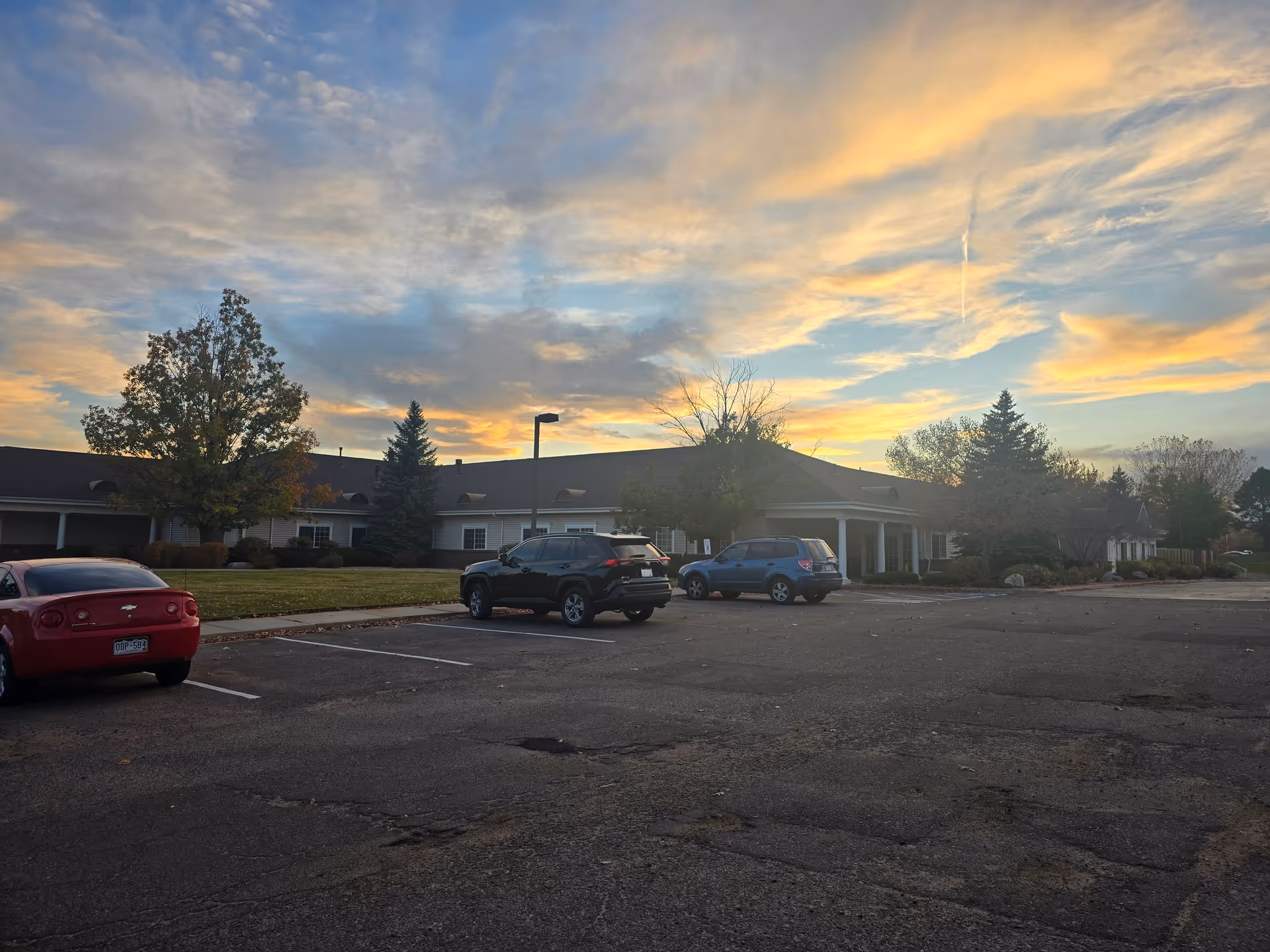 Front exterior of a single-story retirement community building with a few parked cars in the lot under a colorful sunset sky.