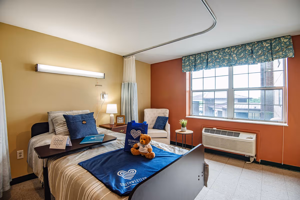 A cozy nursing facility bedroom with a single bed covered in striped bedding and a blue Southwestern Nursing and Rehabilitation Center blanket. A teddy bear and a blue bag with the facility's logo are placed on the bed. Next to the bed is a small wooden nightstand with a lamp and a clock. A white cushioned chair with a blue pillow sits near a large window with floral valance curtains. Below the window is an air conditioning unit.