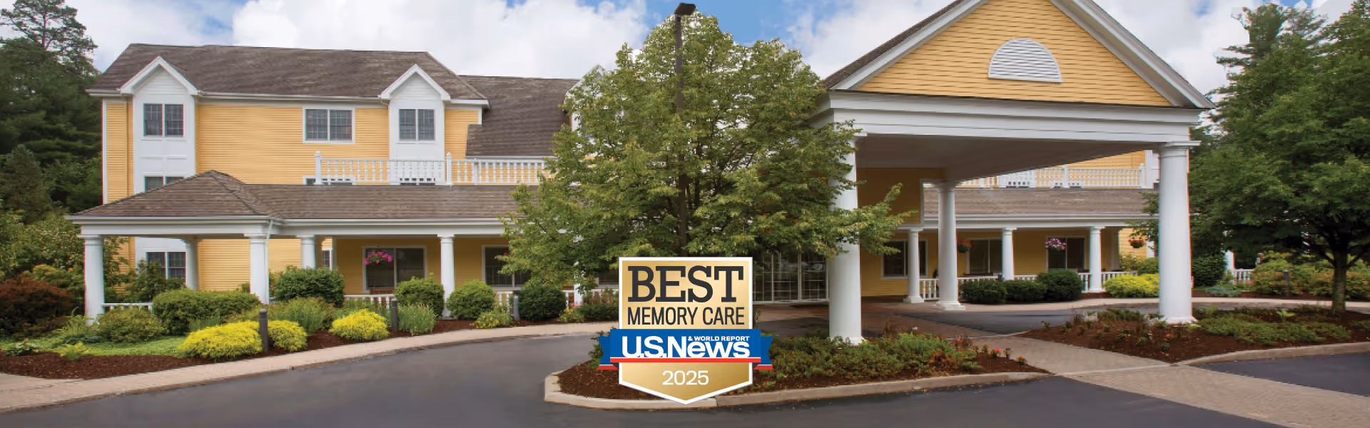 Front exterior view of a large yellow senior living facility building with white trim, a covered entrance supported by white columns, landscaped bushes and trees, and a sign in front that reads 'Best Memory Care U.S. News 2025'.