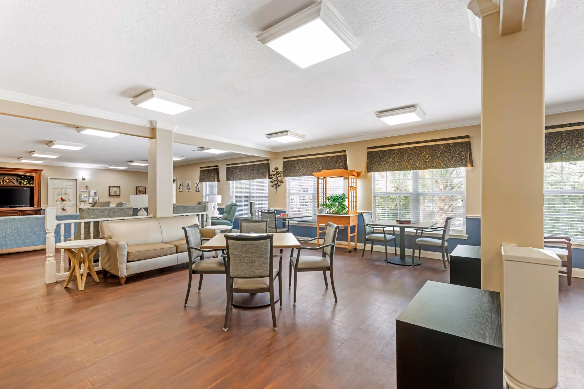 Open communal lounge with sofas, chairs, and dining tables arranged near large windows in a senior living facility.