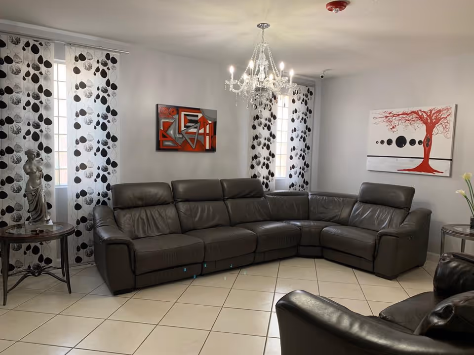 Bright living room featuring a large gray leather sectional under a chandelier, patterned curtains, and wall art.