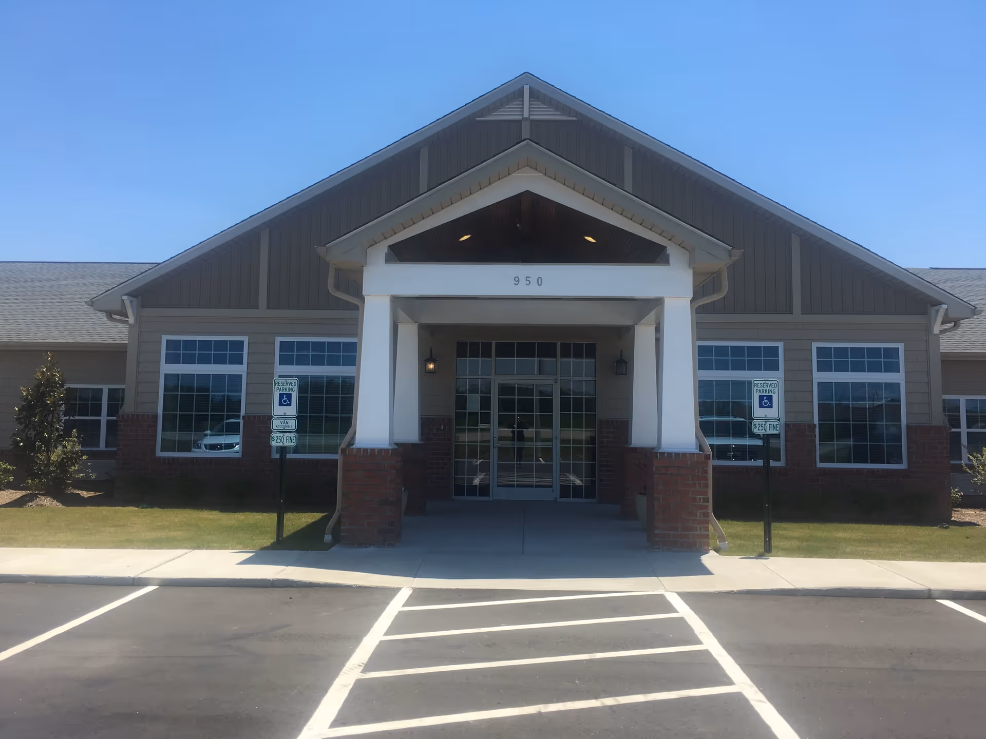 Front exterior view of a single-story building with a peaked roof and large windows. The entrance has a covered porch supported by white columns with brick bases. There are two reserved parking signs for handicapped parking on either side of the entrance. The building number 950 is displayed above the entrance.