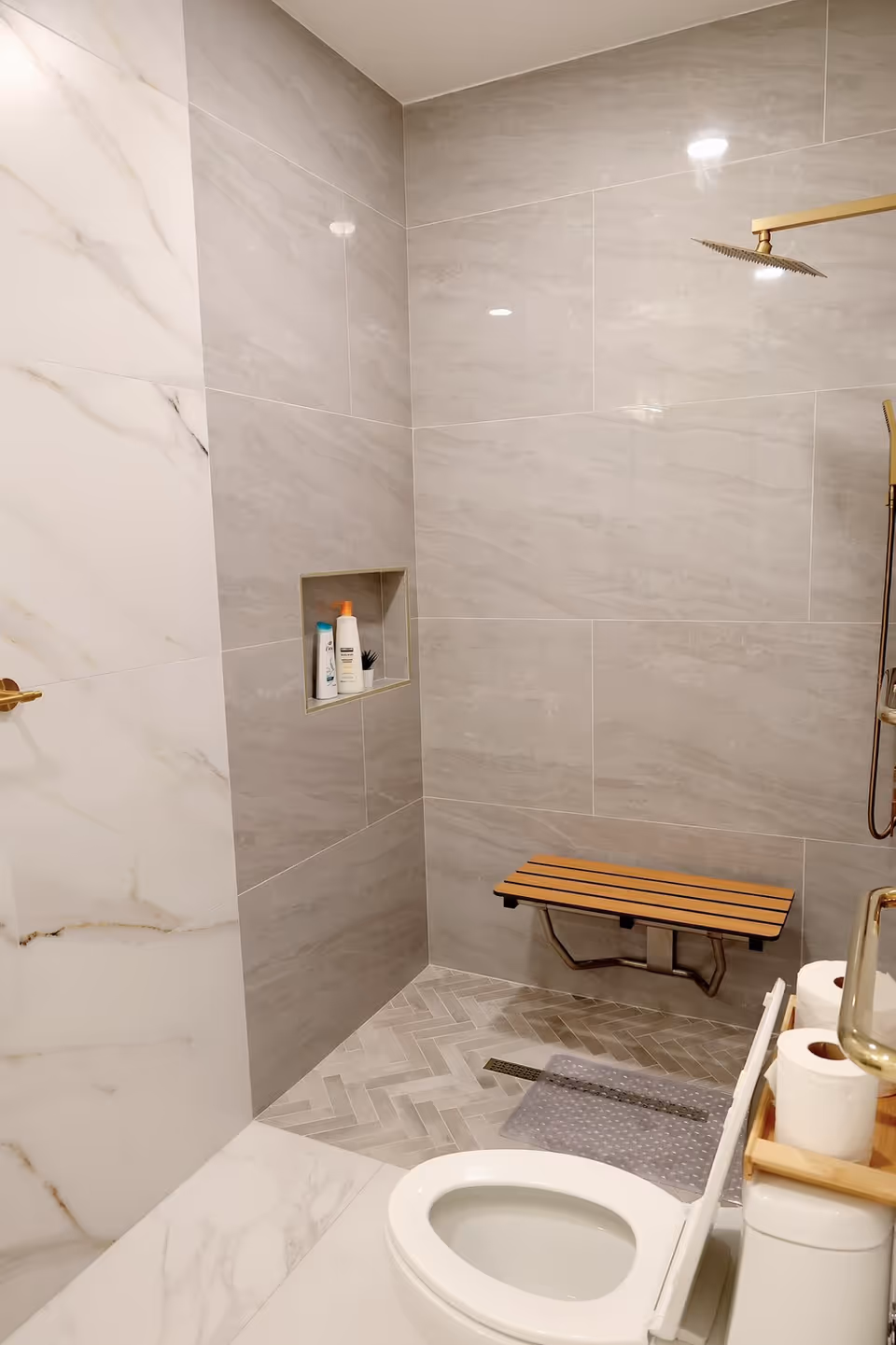Modern bathroom with a walk-in shower featuring large gray and white marble tiles, a built-in niche holding shampoo and lotion bottles, a wooden fold-down shower bench, a rain shower head with gold fixtures, and a white toilet with a wooden shelf holding extra toilet paper rolls.