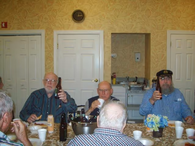 A group of elderly men sitting around a table in a room with patterned wallpaper, drinking from bottles and cups, with plates of food and a flower centerpiece on the table. There are white doors and a clock on the wall behind them.