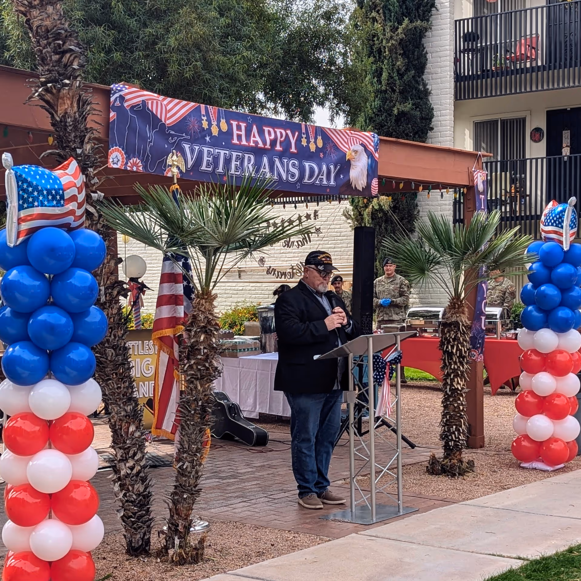An outdoor Veterans Day event at Fellowship Square Tucson with a man speaking at a podium under a decorated pergola. The area is adorned with red, white, and blue balloon columns featuring American flags. Several people in military uniforms stand in the background near tables with food and decorations.