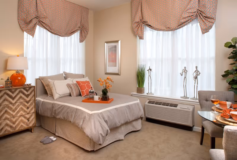 A cozy bedroom in an assisted living facility featuring a neatly made bed with multiple pillows and a tray with a coffee pot and cup. The room has two large windows with patterned valances and sheer white curtains, a wooden nightstand with a lamp and decorative items, a framed picture on the wall, and a small round dining table with chairs set with orange dishes and glasses. There are also decorative metal figurines on the windowsill and a potted plant in the corner.