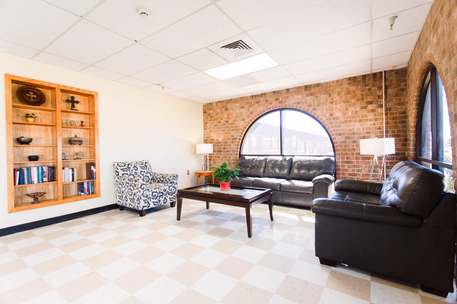 A bright living room with a checkered tile floor, two black leather sofas, a patterned armchair, a wooden coffee table with a potted plant, two floor lamps, a built-in wooden shelf with decorative items and books, and large arched windows with a brick wall background.