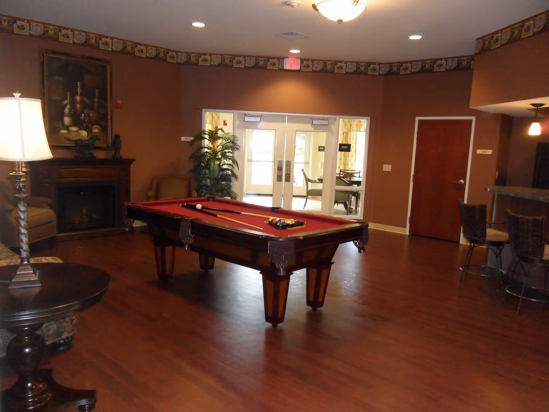 A recreational room with a red felt pool table in the center, two pool cues and a rack of billiard balls on it. The room has wooden flooring, brown walls with a decorative border near the ceiling, a fireplace with a painting above it, a floor lamp, and some chairs. There is a double glass door leading to another room with tables and chairs, and a bar area with two bar stools on the right side.