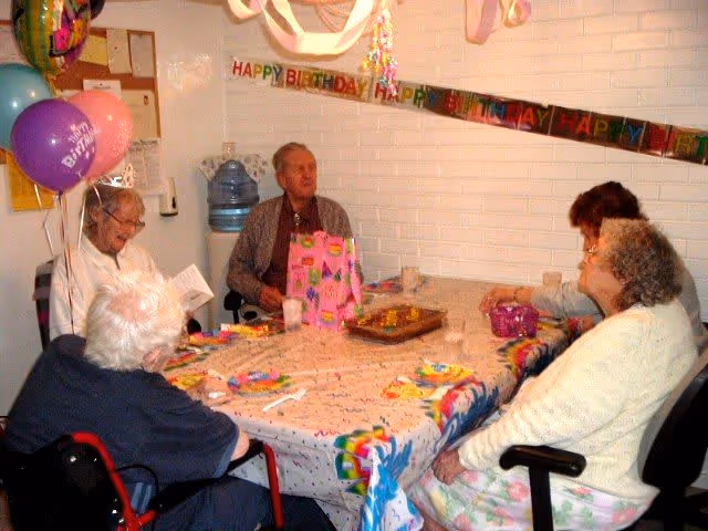 Five elderly residents seated around a decorated table with a birthday cake, balloons, and a gift in a communal room.