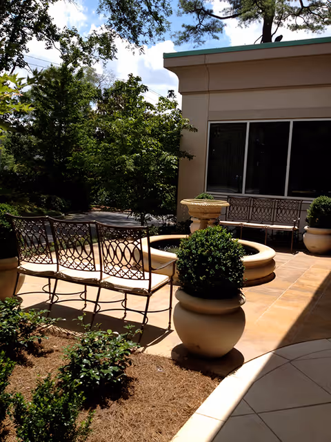 Outdoor patio area with metal benches and a circular stone fountain surrounded by potted plants and greenery, adjacent to a building with large windows under a partly cloudy sky.