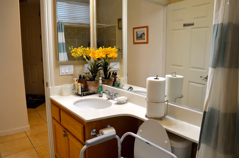 A bathroom with a white countertop and wooden cabinets. On the countertop, there are two paper towel rolls, a soap dispenser, a bar of soap, and a decorative plant with yellow flowers. A large mirror reflects the room, showing a door labeled 'Restrooms' and a shower curtain with blue and white stripes. The floor is tiled, and there is a handrail attached to the toilet for support.