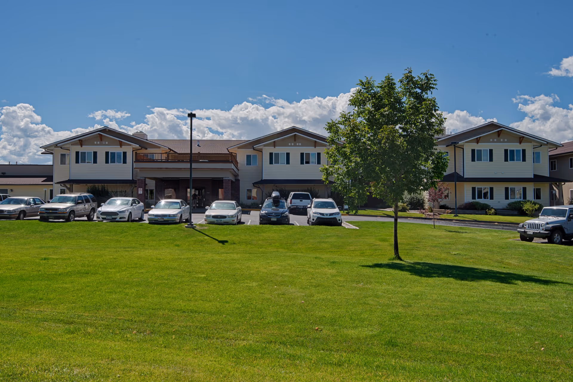 Front exterior of a two-story senior living building with a wide green lawn, a row of parked cars, and a tree under a blue sky.