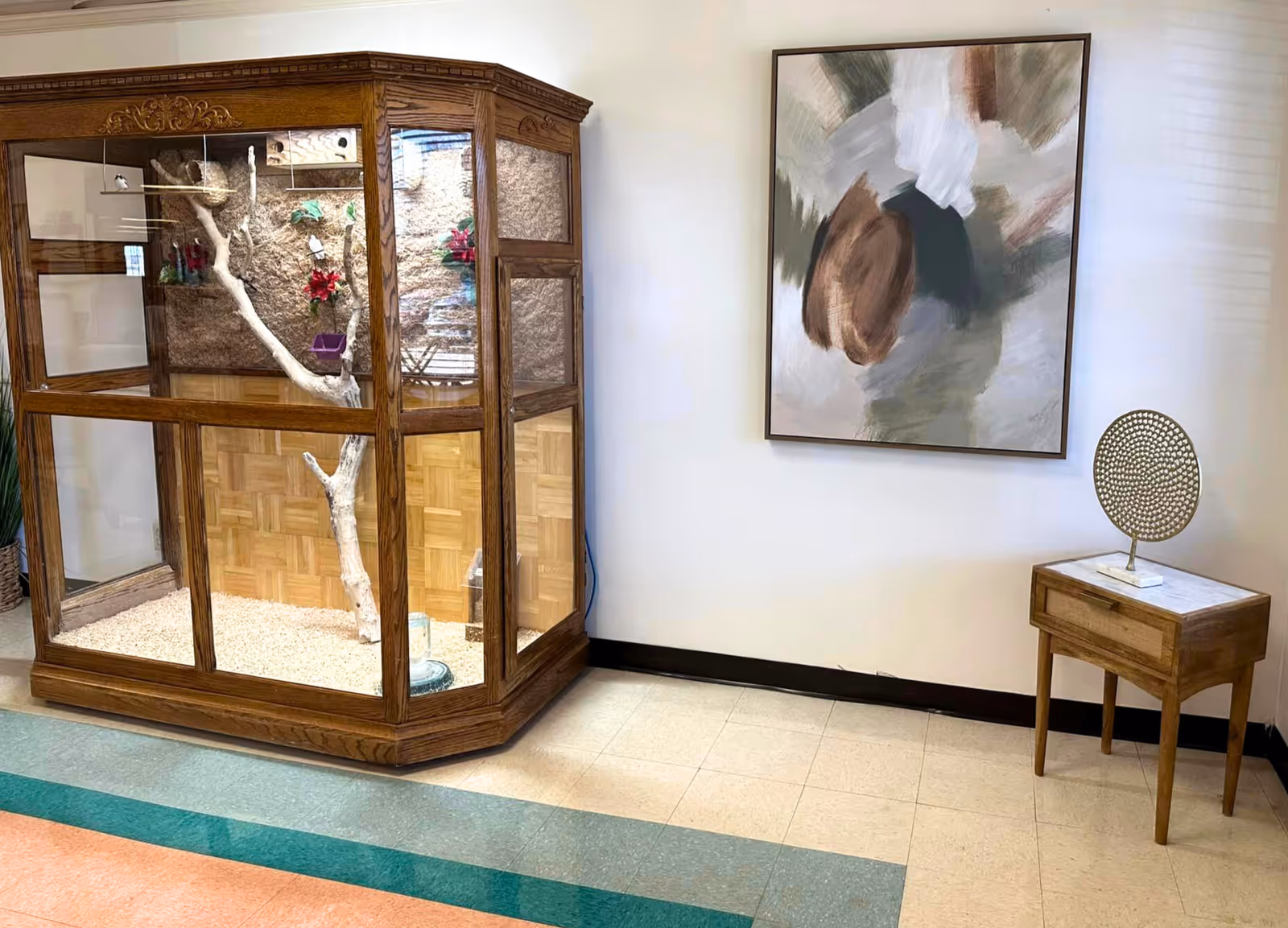 An indoor corner of a facility featuring a large wooden and glass enclosure with a tree branch inside, possibly for birds or small animals. Next to it is a small wooden side table with a decorative circular sculpture on top. A large abstract painting with earthy tones hangs on the white wall behind the table.