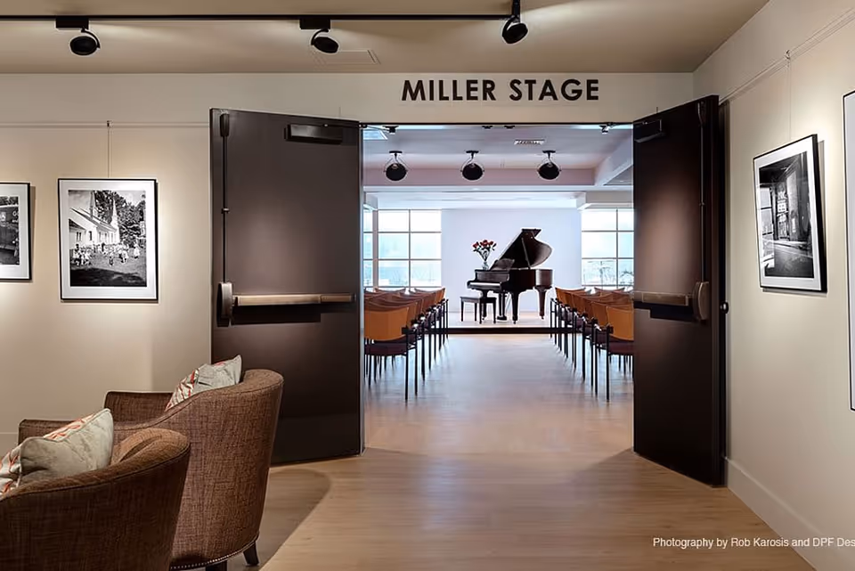 View through open double doors labeled 'Miller Stage' into a small auditorium with rows of chairs facing a grand piano on a stage. The foreground shows a cozy seating area with two brown armchairs and framed black-and-white photographs on the walls.