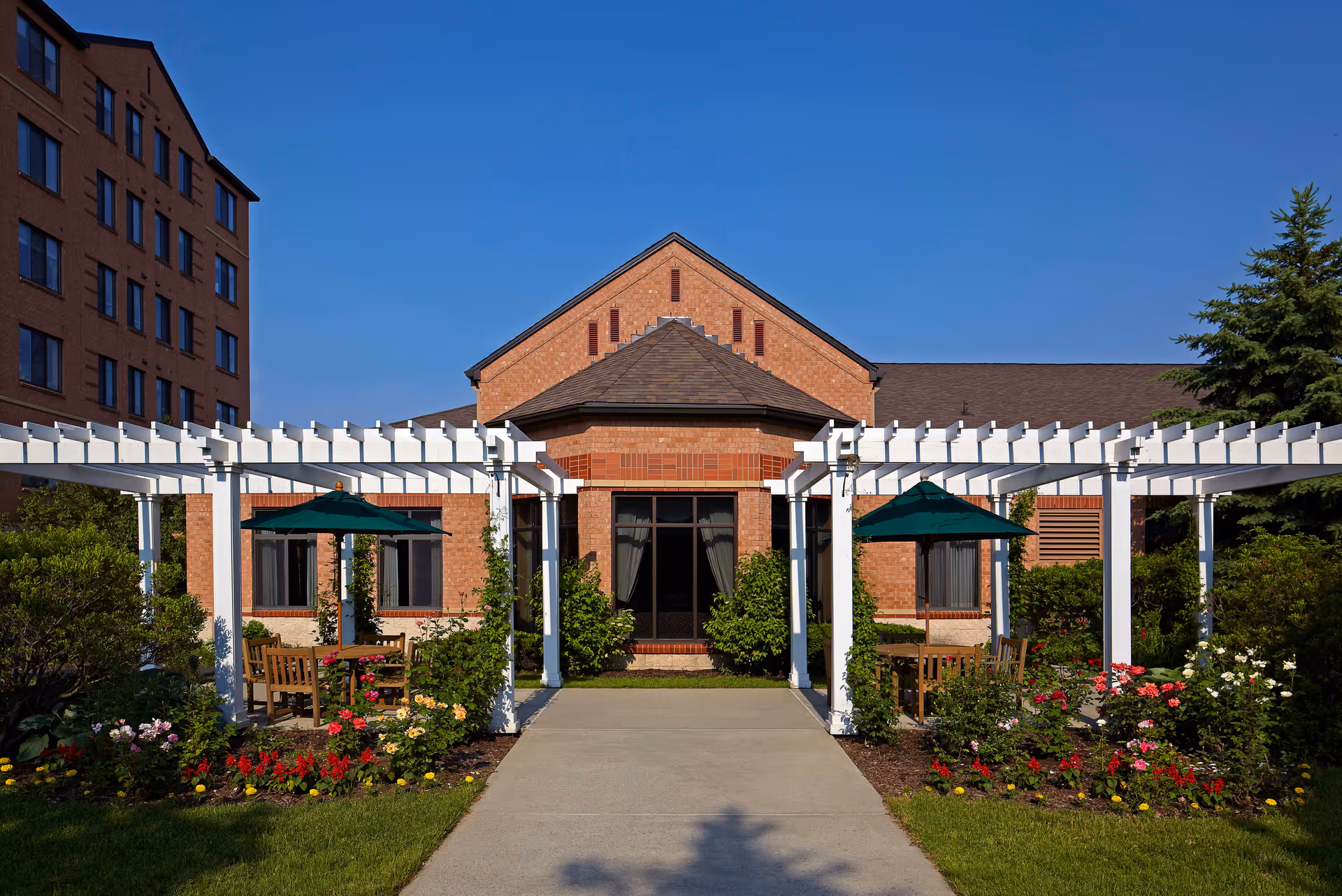 Outdoor garden area at Allegria Village featuring a paved walkway leading to a brick building with large windows. White pergolas with climbing plants frame the walkway, and there are wooden tables with green umbrellas surrounded by colorful flower beds and greenery under a clear blue sky.