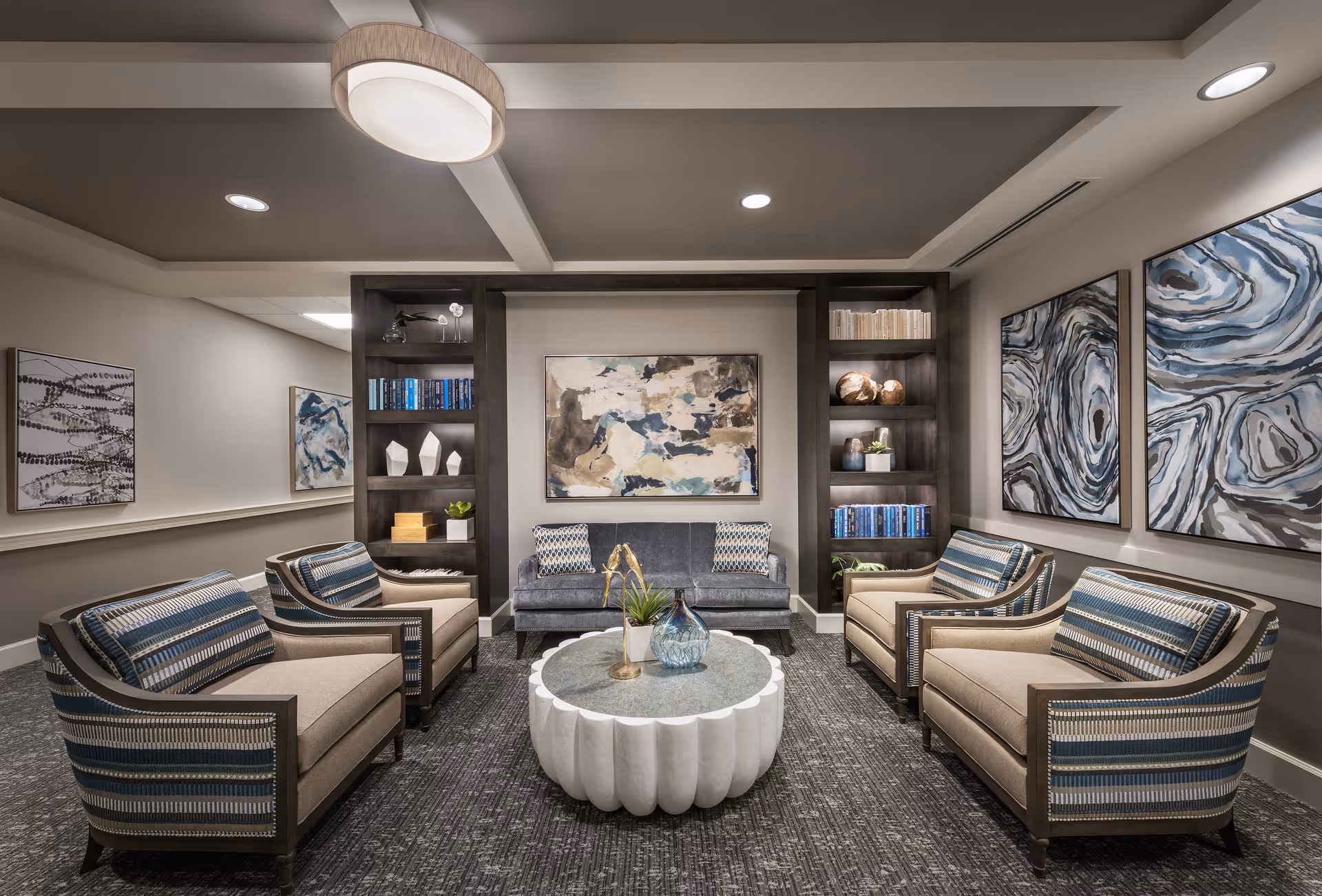A modern and cozy living room area with four beige and blue striped armchairs arranged around a white scalloped round coffee table with decorative items on top. A gray sofa with patterned cushions is against the back wall, flanked by built-in dark wood shelves filled with books and decorative objects. Abstract artwork in neutral and blue tones adorns the walls, and the room is softly lit by ceiling lights and a central pendant light.