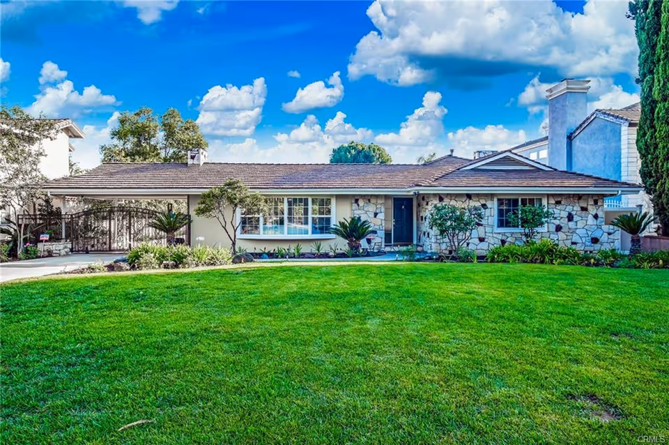 Single-story residential building with a stone facade and a large green lawn in front under a bright blue sky with scattered clouds. The house has multiple windows and a dark front door, surrounded by small shrubs and plants.