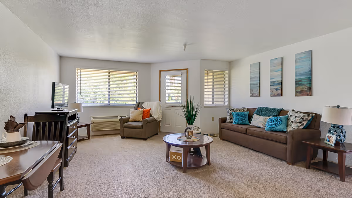 A bright and cozy living room in a senior living community featuring a brown sofa with multiple decorative pillows, a matching armchair with a throw blanket, a round wooden coffee table with a plant centerpiece, a wooden side table with a lamp and framed photo, a TV on a stand, and large windows with blinds letting in natural light.