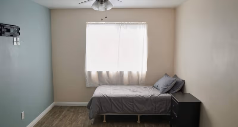 A small bedroom with a single bed covered in gray bedding and two pillows. There is a black nightstand with drawers next to the bed. A window with sheer white curtains lets in natural light. The walls are painted beige and one wall is painted teal. A ceiling fan with lights is mounted on the ceiling. The floor has wood-like flooring.