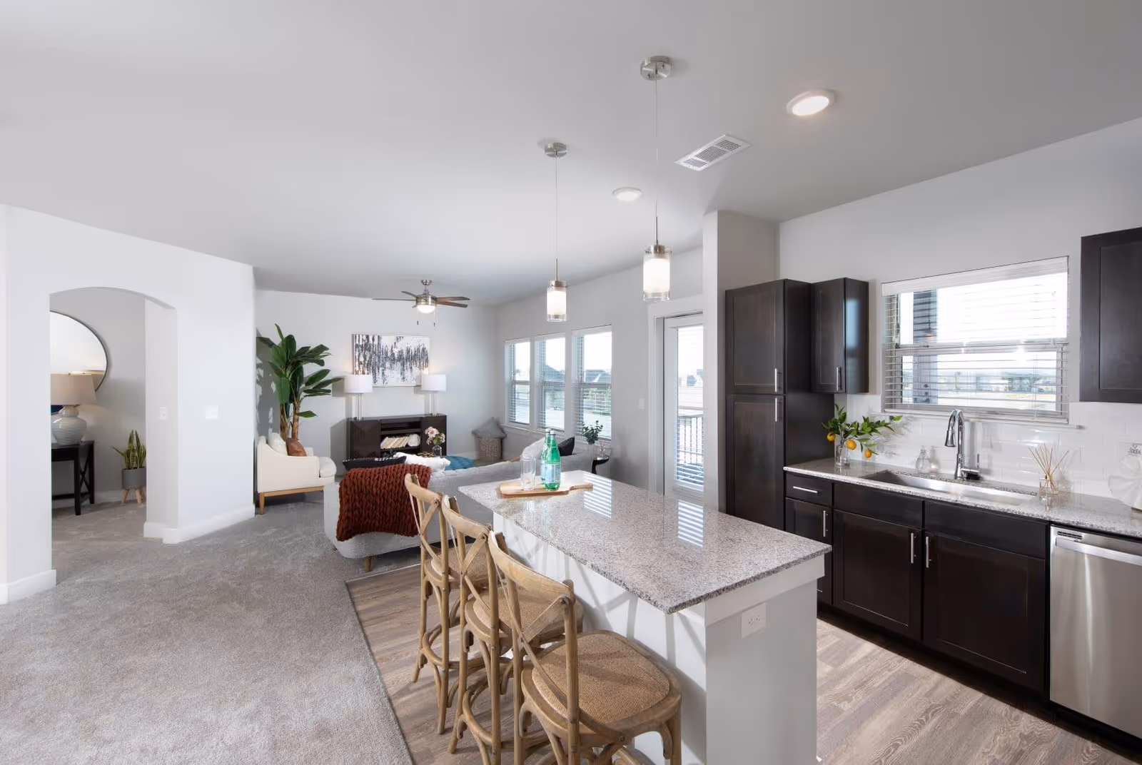Bright and modern open-concept living space featuring a kitchen with dark wood cabinets, a granite countertop island with three wooden bar stools, and a living room area with a sofa, armchair, plants, and wall art. Large windows and a glass door allow natural light to fill the room.