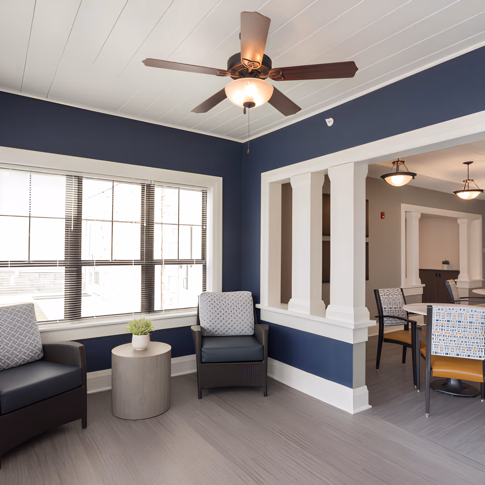 A cozy seating area in a senior living facility with two cushioned armchairs and a small round table with a potted plant between them. The walls are painted dark blue with white trim, and a ceiling fan with a light fixture is mounted above. Adjacent to the seating area is a dining space with tables and chairs, separated by white columns and partial walls.