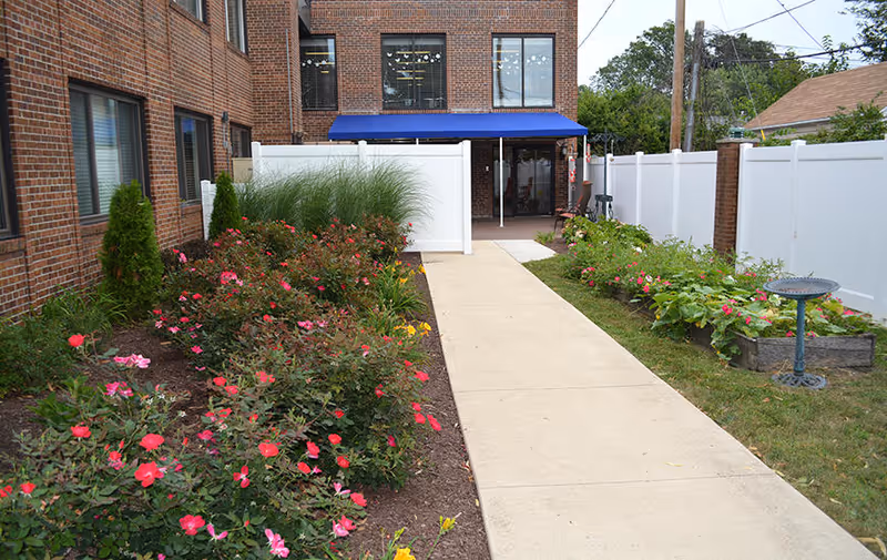 A concrete walkway leads through a garden area with blooming pink and red flowers on the left and green plants on the right, bordered by a white fence. The walkway leads to the entrance of a brick building with a blue awning above the door.