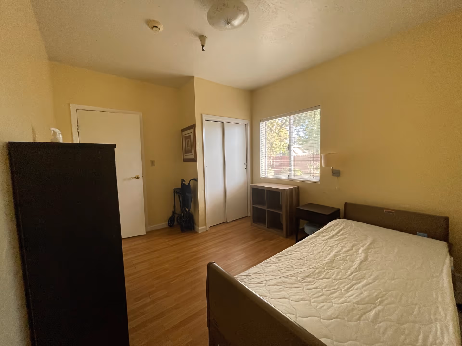 A simple bedroom with a bed covered by a white mattress, a small nightstand, a wooden shelf unit under a window with blinds, a closed closet with white sliding doors, a door, and a walker placed in the corner. The room has light yellow walls and wooden flooring.
