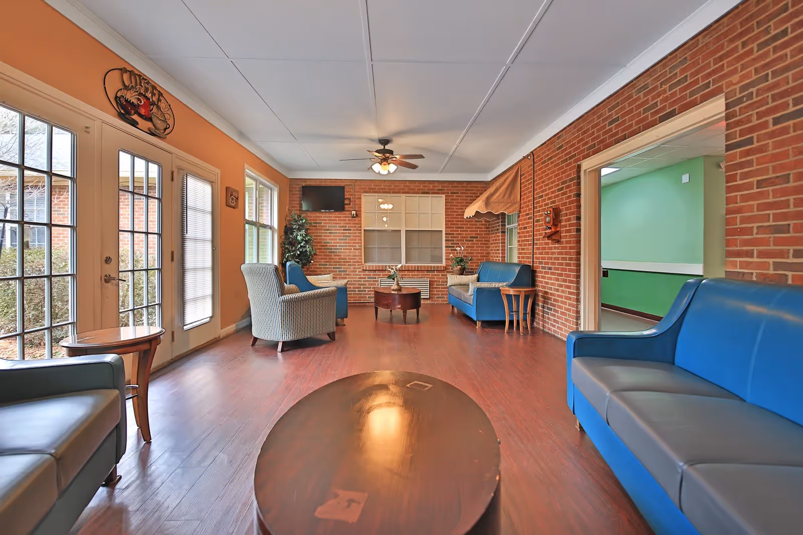 A cozy indoor seating area with blue sofas and striped armchairs arranged around wooden coffee tables. The room features brick walls, large windows, a ceiling fan with lights, and a door leading outside. There is a decorative sign above the door that says 'Coffee'.