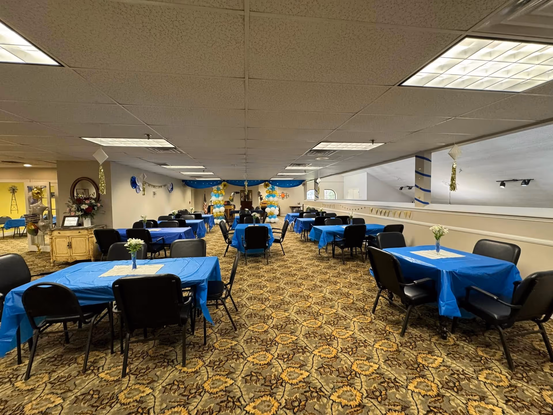 A large room set up for an event with multiple tables covered in blue tablecloths and black chairs around them. Each table has a small vase with flowers. The room has patterned carpet, a drop ceiling with fluorescent lights, and decorations including balloons and banners at the far end near a podium.