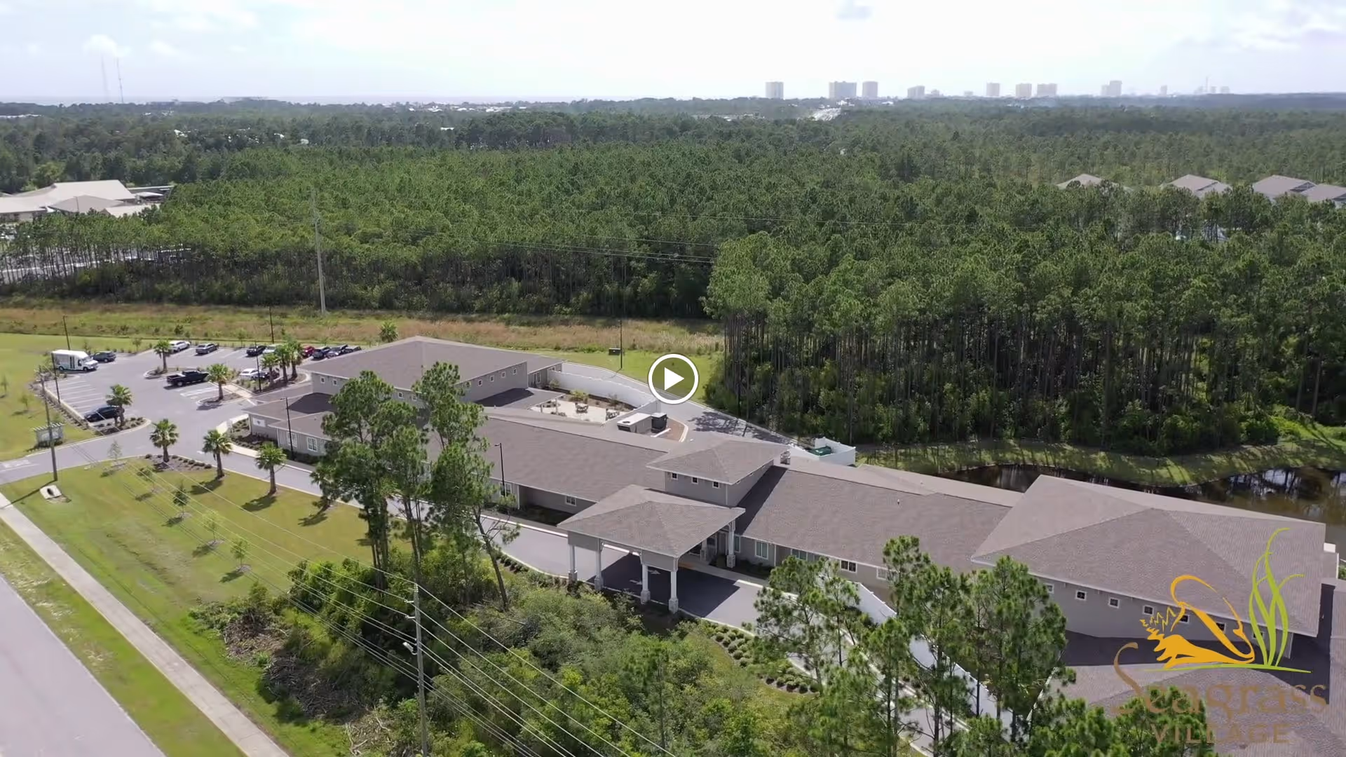 Aerial view of The Landing of Panama City Beach senior living facility surrounded by greenery and trees, with a parking lot and a road nearby. The building has a large, single-story layout with a covered entrance and multiple roof sections.