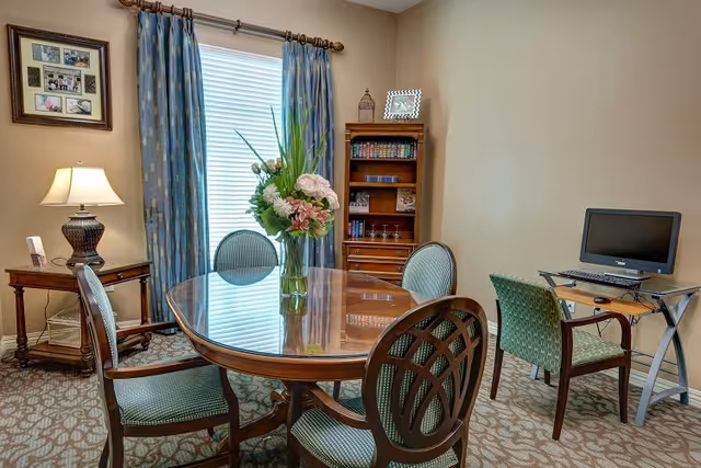 A cozy room with a polished wooden oval table surrounded by four upholstered chairs. A vase with flowers sits on the table. To the left, there is a wooden side table with a lamp and framed photos on the wall above it. In the background, a window with blue patterned curtains lets in natural light. On the right side, there is a small desk with a computer monitor and a chair. A wooden bookshelf with books and decorative items is against the wall.