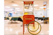 Interior view of a large dining area with multiple tables covered in green and yellow tablecloths. In the foreground, there is a vintage-style red popcorn machine with yellow wheels. The room is brightly lit with white walls and ceiling lights.