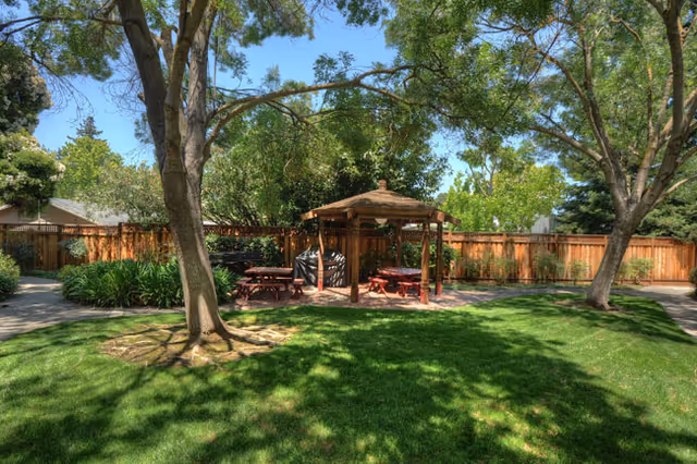 Shaded grassy courtyard with large trees, a wooden gazebo surrounded by picnic tables, and a wooden fence.