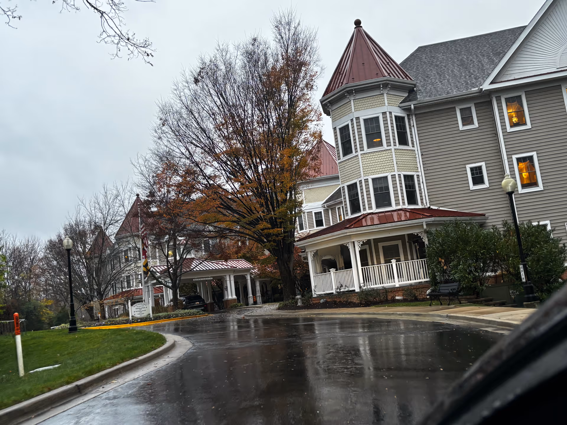 Exterior view of a senior living facility building with gray siding, white trim, and red metal roofs on turret-like structures. The scene is on a rainy day with wet pavement, leafless trees, and some autumn foliage. There is a curved driveway leading to a covered entrance with a sign and an American flag nearby.