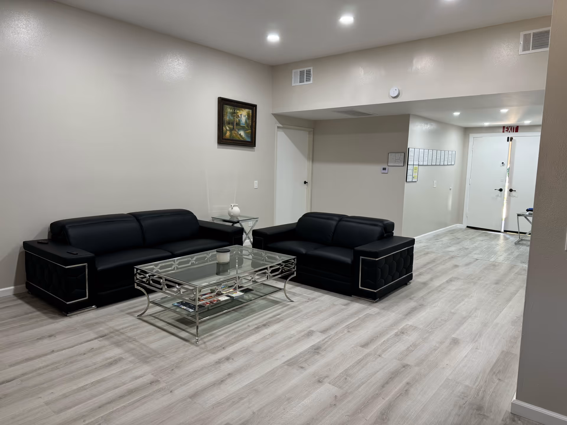 A minimal common lounge with two black leather sofas, a glass coffee table, light wood floors and a doorway to an exit.