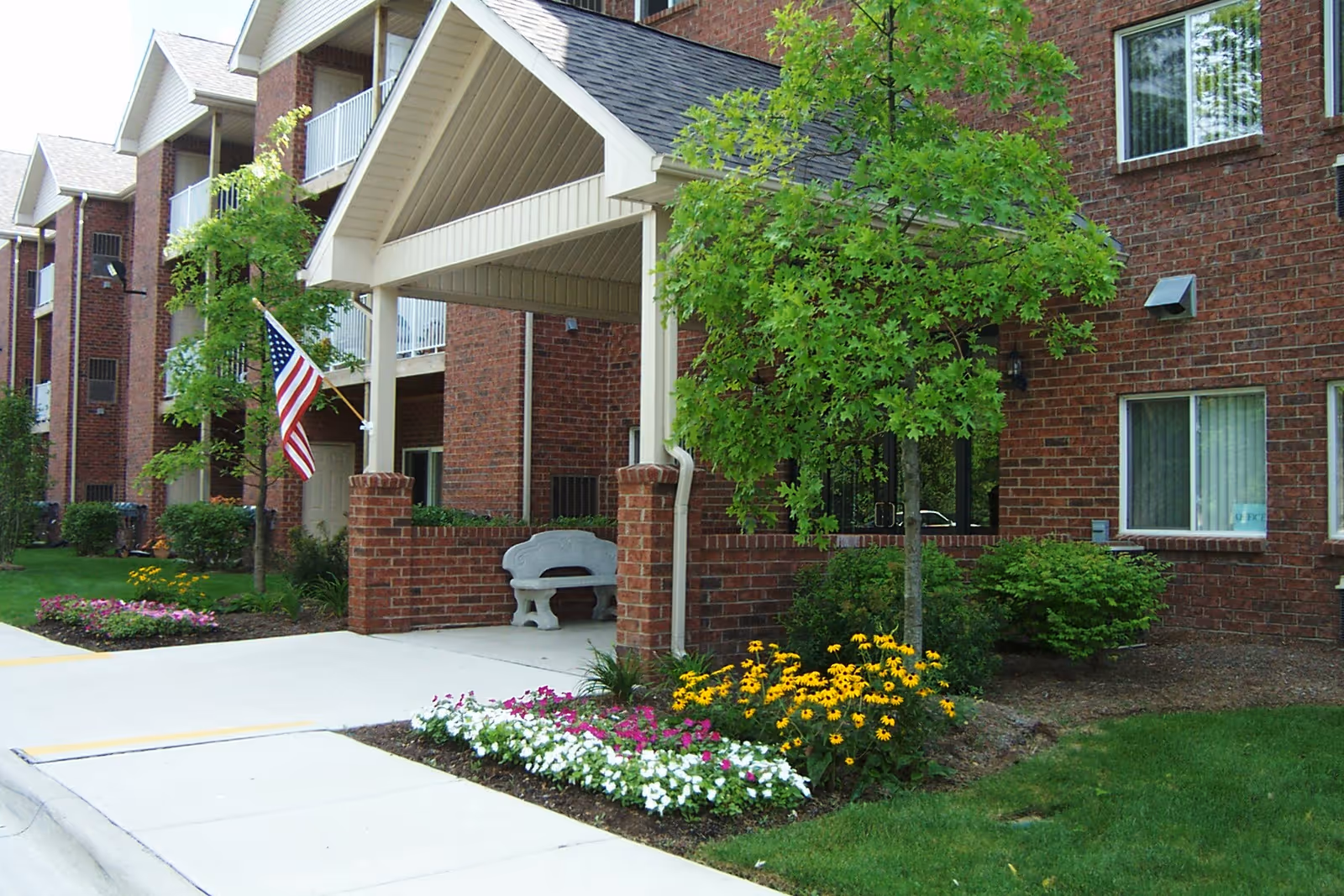 Covered brick entrance of a senior living building with an American flag, bench, and flower beds.