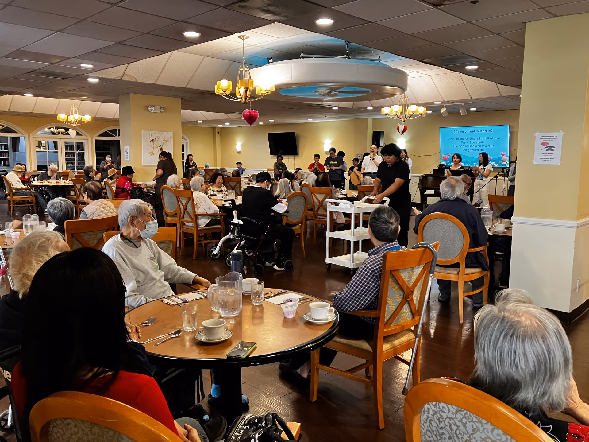A large dining room filled with elderly residents seated at round tables, some wearing masks. A group of people is performing music at the front of the room near a piano and a screen displaying song lyrics. The room is warmly lit with chandeliers and decorated with hanging heart ornaments.
