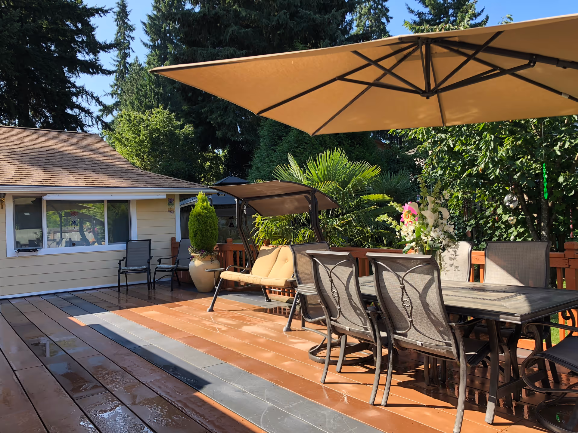 Outdoor patio area with a large beige umbrella providing shade over a dining table with six chairs. There is a cushioned swing seat nearby, potted plants, and lush green trees surrounding the space. A building with a window and two chairs is visible in the background.