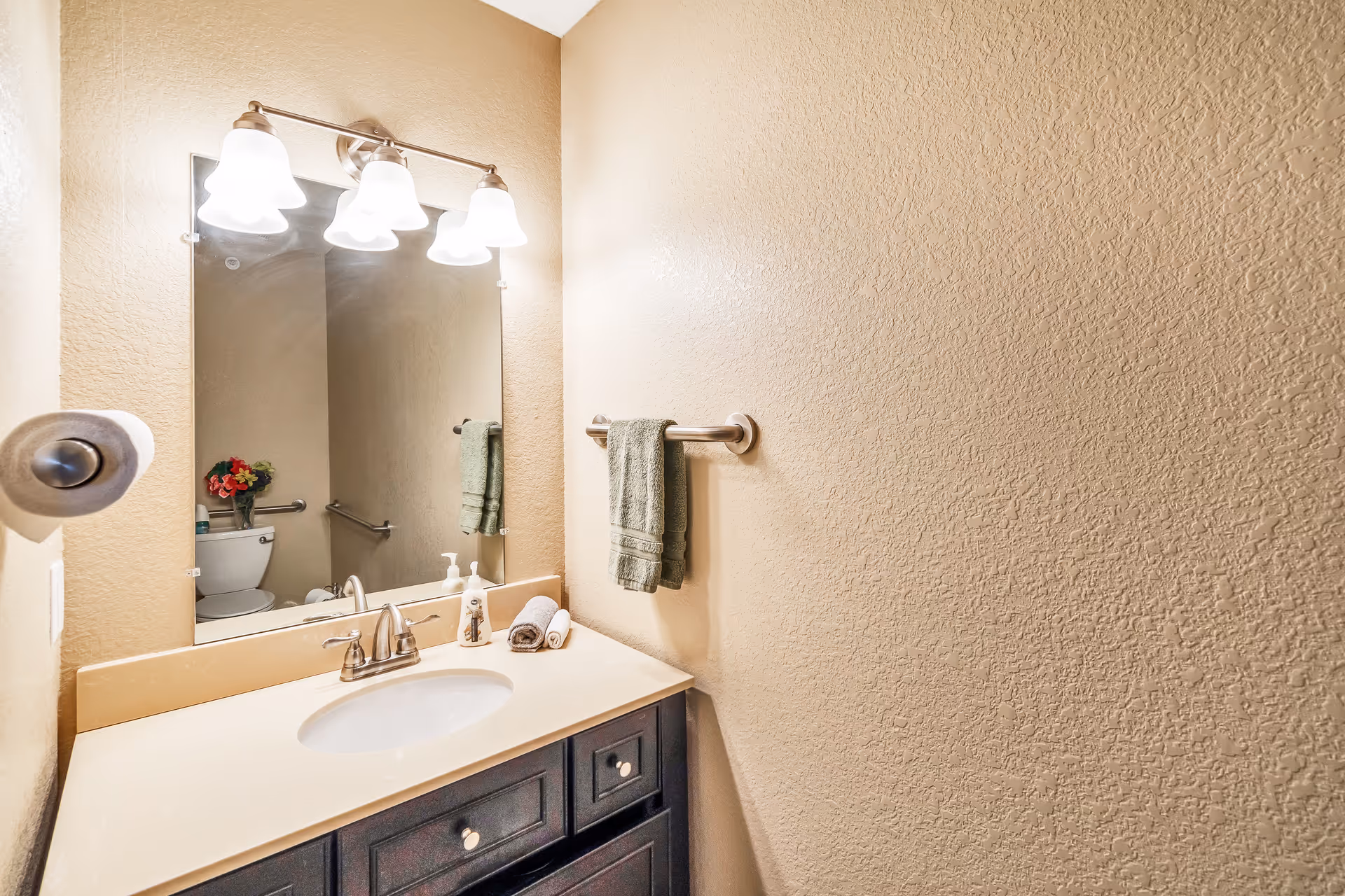 A small bathroom with a beige textured wall, a sink with a white countertop and dark wooden cabinet below. Above the sink is a large mirror with a three-light fixture. A green hand towel hangs on a towel bar next to the sink, and a toilet with a grab bar and a small vase of flowers on the tank is visible in the mirror reflection.