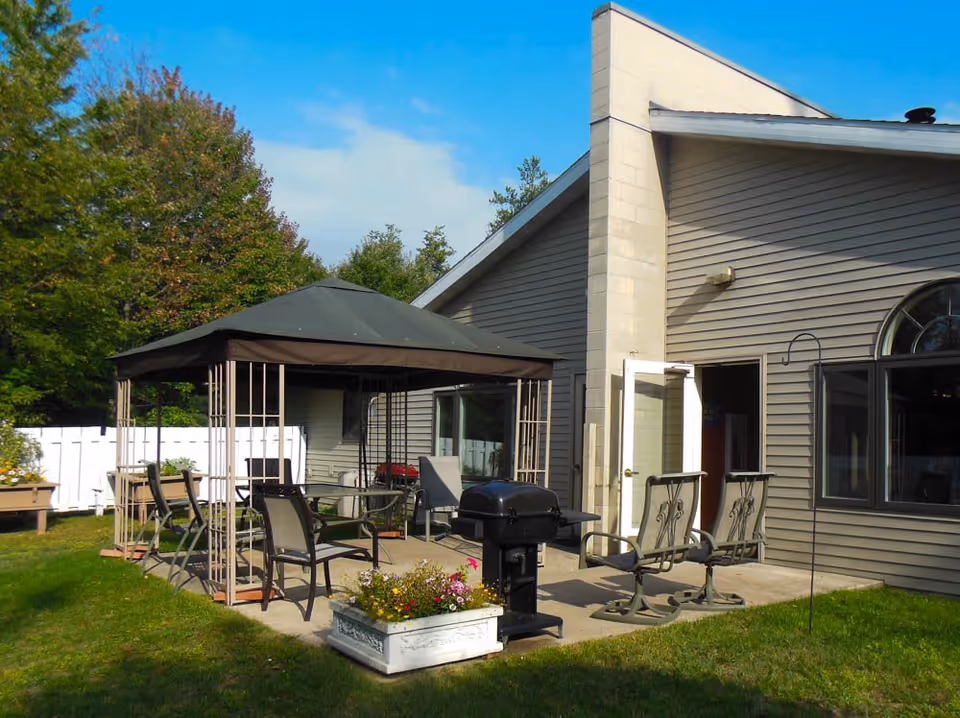 Outdoor patio area at Sylvan Crossings of Stevens Point featuring a gazebo with several chairs underneath, a black grill, two metal chairs with armrests, a flower planter with colorful flowers, and a building with beige siding and large windows in the background.