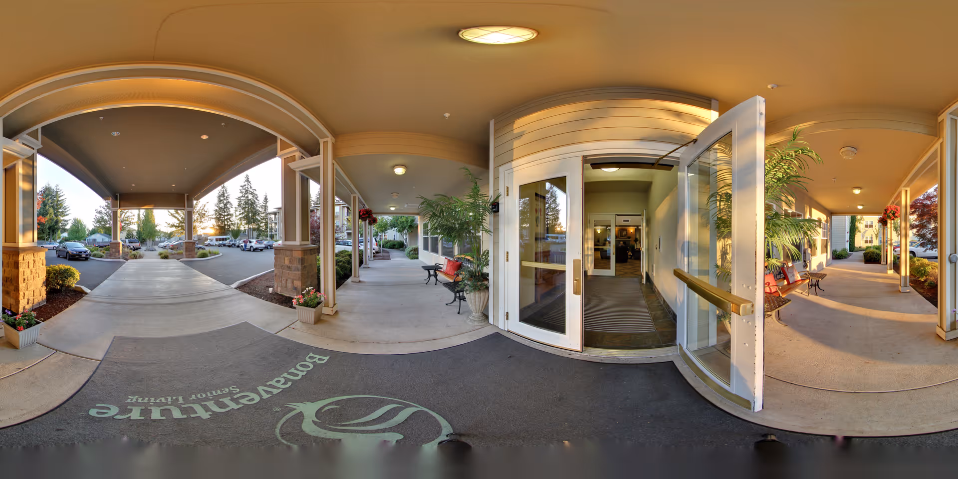 Covered entrance area of Silver Creek by Bonaventure senior living facility with open glass doors leading inside. There are benches, potted plants, and a driveway with parked cars visible outside under a clear sky.