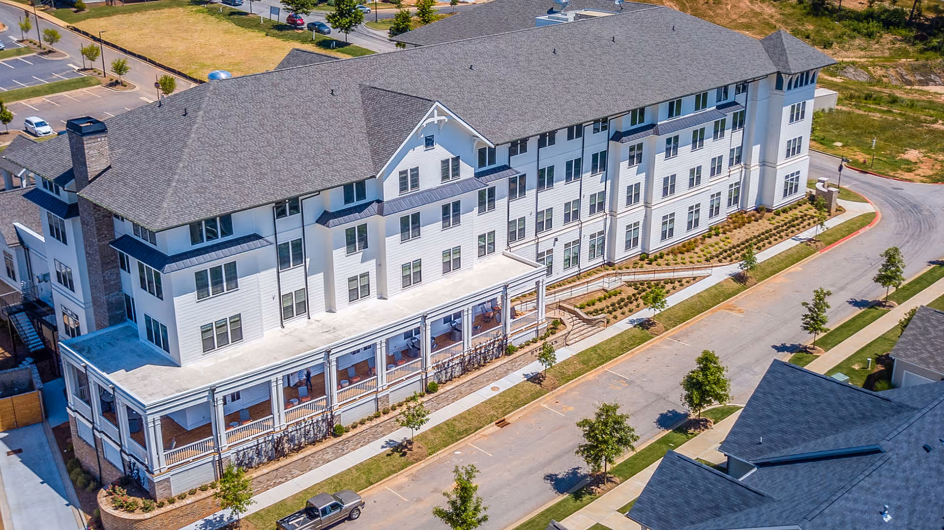 Aerial view of a multi-story white senior living building with a covered wraparound porch, landscaped grounds, and an adjacent road.