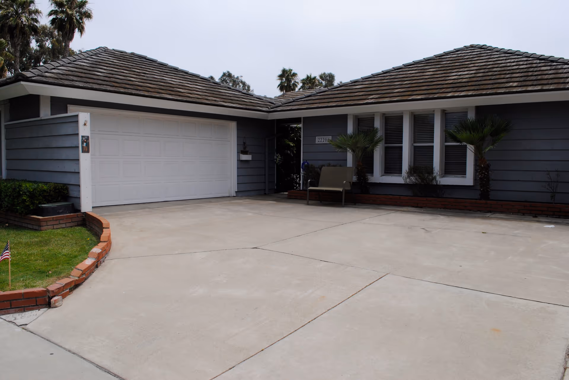 Exterior view of a single-story house with a gray roof and blue siding. The house features a large white garage door, a concrete driveway, a small patch of grass with a brick border, two small palm trees near the windows, and a bench near the entrance.